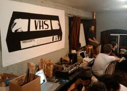 A photo of the interior of VHS taken some time in 2009. A large poster on the wall shows an older VHS logo, and you can see a few people working and talking in the space
